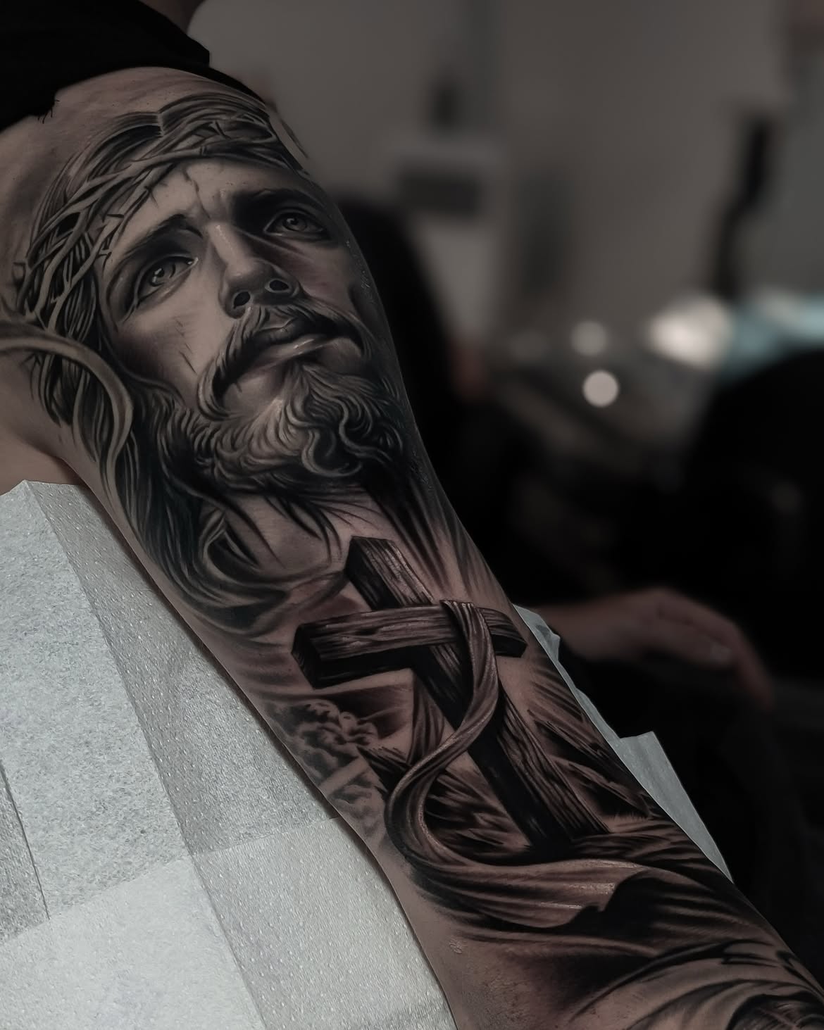 Realistic black and grey tattoo of Jesus Christ with a cross on the upper arm by Alejandro 